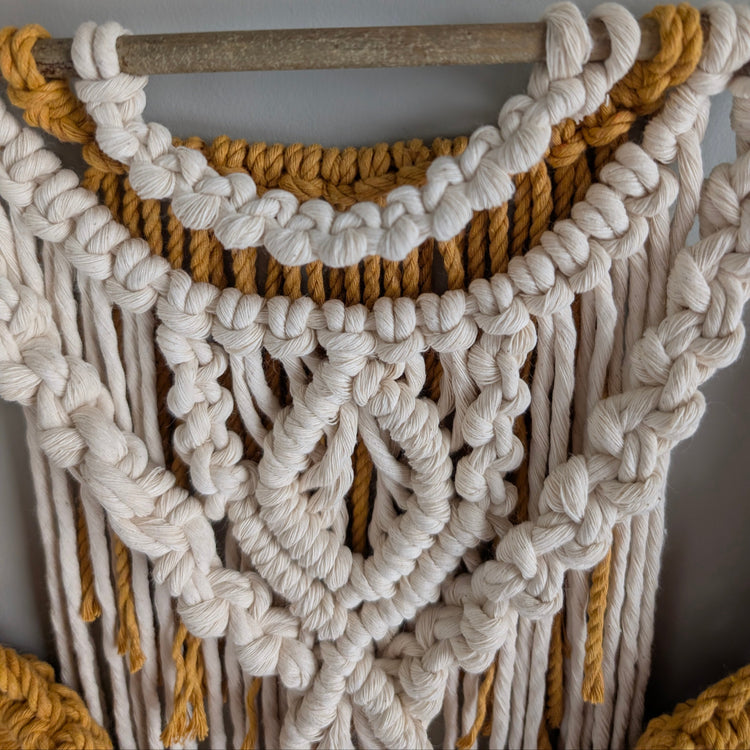Macrame wall hanging with white and gold ropes against a neutral background