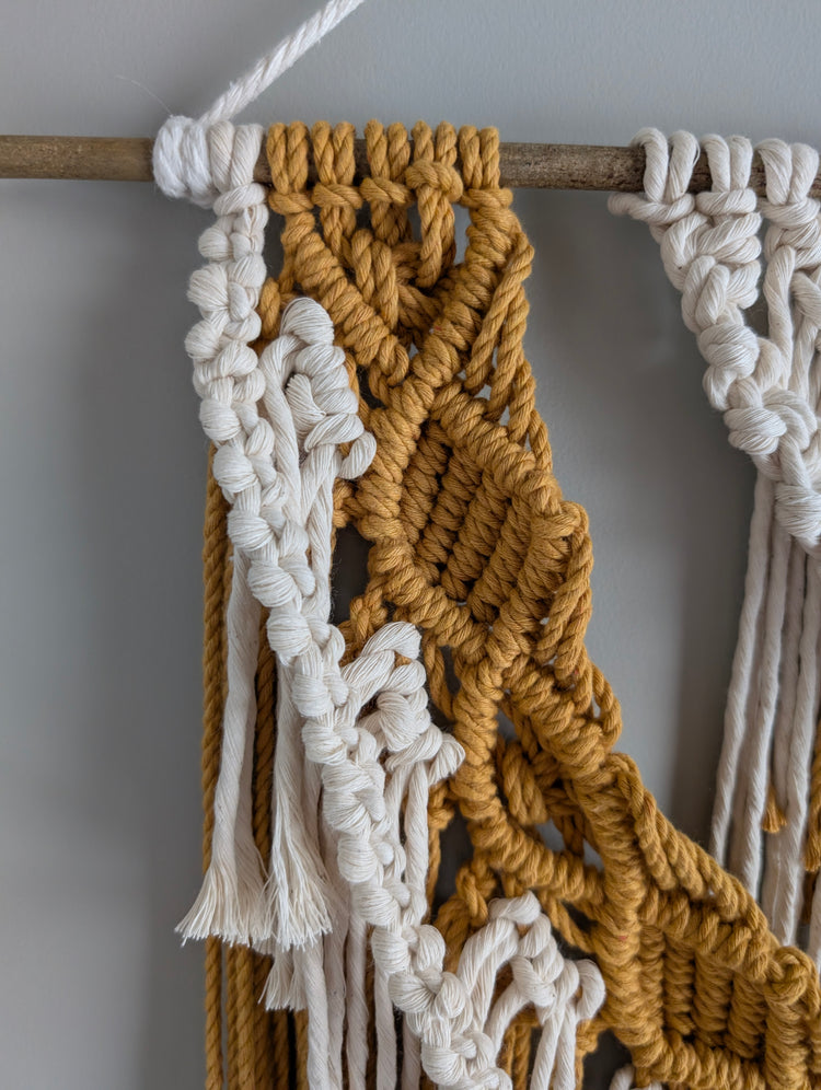 Close-up of a macrame wall hanging with white and mustard ropes on a plain background