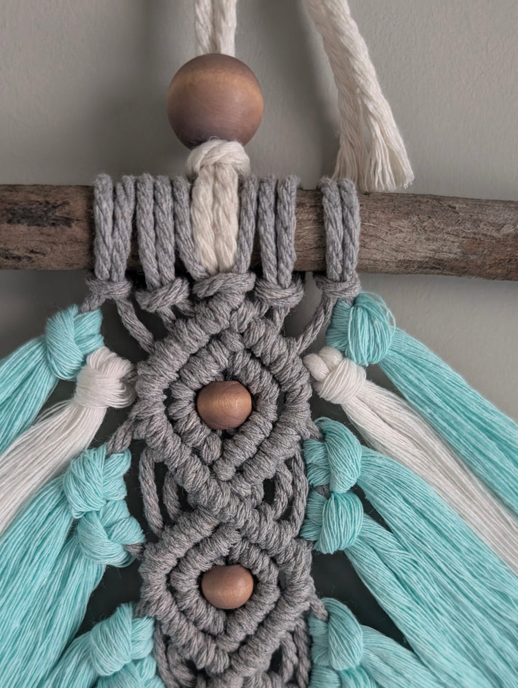 Close-up of a macrame wall hanging with teal, gray, and white ropes and wooden beads.