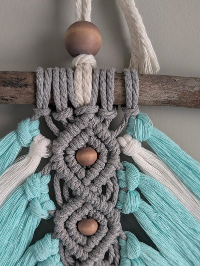 Close-up of a macrame wall hanging with teal, gray, and white ropes and wooden beads.